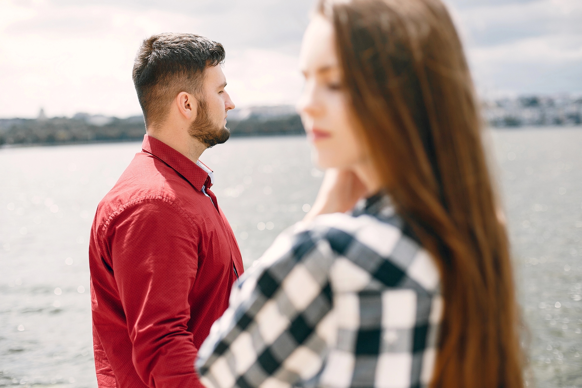 A man in a red shirt looks thoughtfully at a body of water while a woman in a checkered shirt stands in the foreground, slightly out of focus. The scene conveys a sense of contemplation near a waterfront.