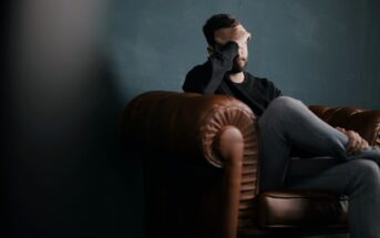 A man sits on a brown leather couch with his legs crossed, resting his elbow on the armrest and covering his eyes with his hand. The background is a dark, muted wall, creating a somber atmosphere.