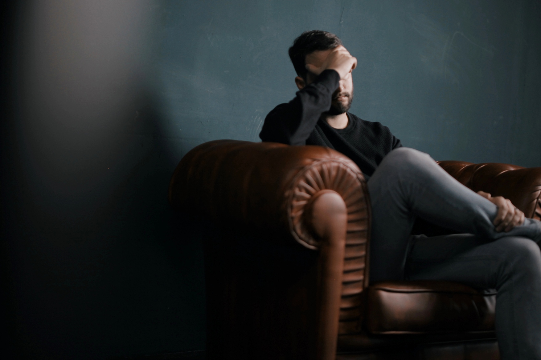 A man sits on a brown leather couch with his legs crossed, resting his elbow on the armrest and covering his eyes with his hand. The background is a dark, muted wall, creating a somber atmosphere.