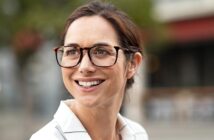 A woman with brown hair and glasses is smiling. She is wearing a white shirt with thin black stripes. The background is softly blurred, featuring hints of greenery and an outdoor seating area.