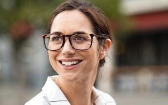 A woman with brown hair and glasses is smiling. She is wearing a white shirt with thin black stripes. The background is softly blurred, featuring hints of greenery and an outdoor seating area.