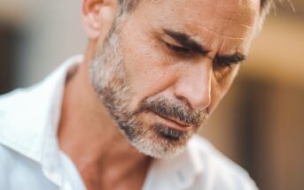 A man with short hair and a beard is intently looking downward. He is wearing a white shirt, and the background is softly blurred.