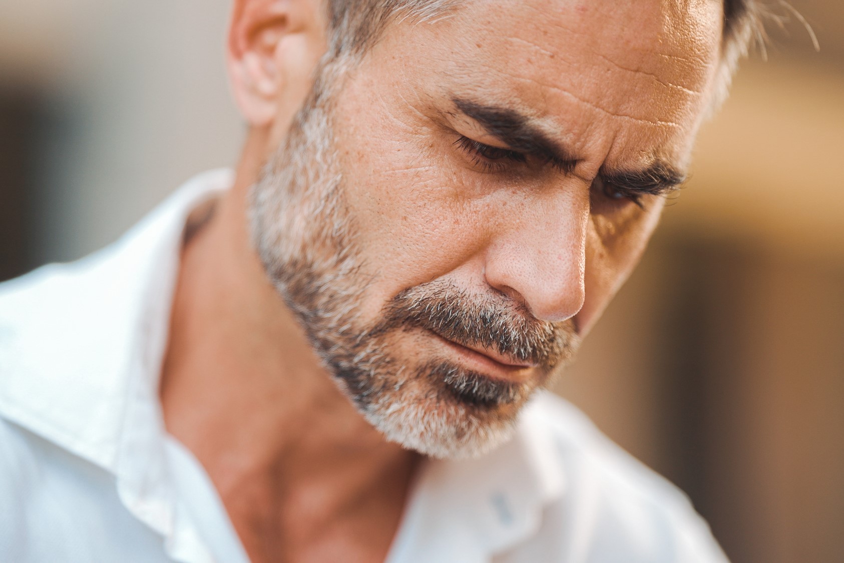 A man with short hair and a beard is intently looking downward. He is wearing a white shirt, and the background is softly blurred.