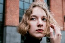 A person with short, wavy blonde hair looks off to the side while standing in front of a brick building. They are touching their hair gently.