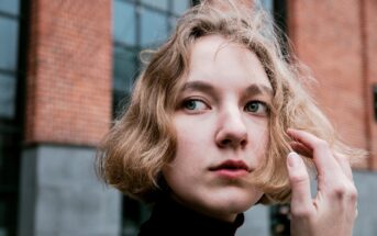 A person with short, wavy blonde hair looks off to the side while standing in front of a brick building. They are touching their hair gently.