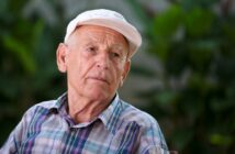 An elderly man wearing a white cap and plaid shirt sits outdoors, looking contemplative. The blurred greenery in the background suggests a garden or park setting.