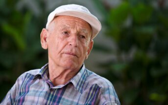 An elderly man wearing a white cap and plaid shirt sits outdoors, looking contemplative. The blurred greenery in the background suggests a garden or park setting.