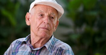 An elderly man wearing a white cap and plaid shirt sits outdoors, looking contemplative. The blurred greenery in the background suggests a garden or park setting.