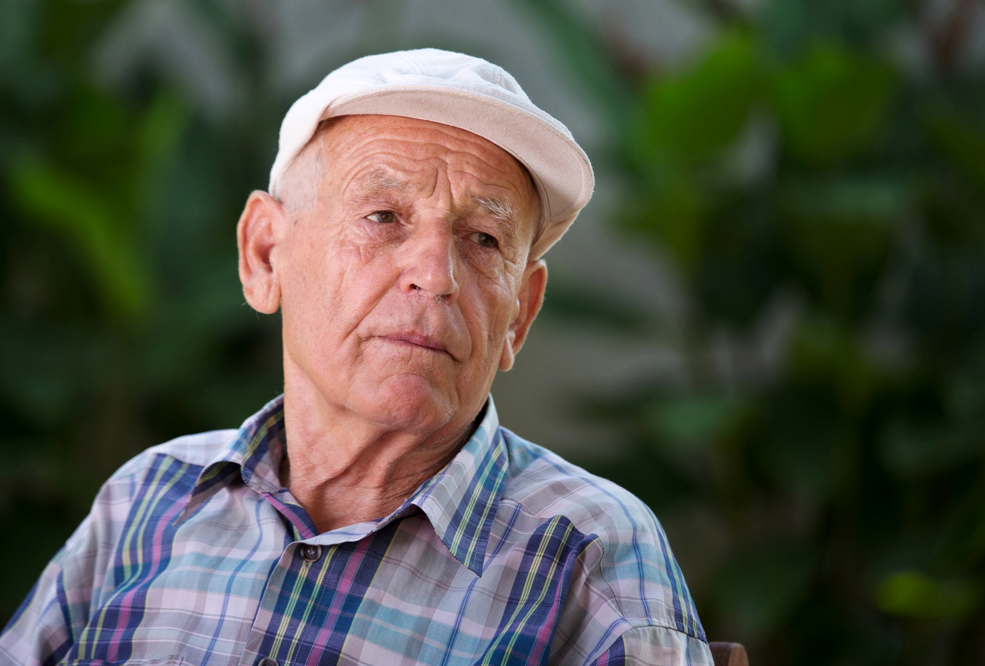 An elderly man wearing a white cap and plaid shirt sits outdoors, looking contemplative. The blurred greenery in the background suggests a garden or park setting.