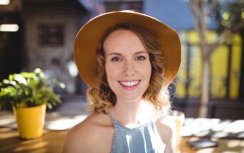 A woman wearing a wide-brimmed brown hat and a light blue sleeveless top is smiling at the camera. She is sitting in an outdoor setting with sunlight casting shadows and a yellow potted plant in the background.