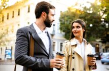 A man and woman walk together on a sunny street, both holding coffee cups. The man is dressed in a suit and carrying a bag, while the woman wears a beige trench coat. Trees and buildings are visible in the background.