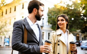 A man and woman walk together on a sunny street, both holding coffee cups. The man is dressed in a suit and carrying a bag, while the woman wears a beige trench coat. Trees and buildings are visible in the background.