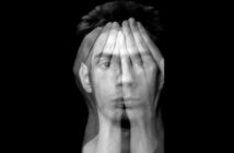 A black and white abstract image showing a man's face blended with overlaying hands covering his eyes. The image creates a double exposure effect, symbolizing vulnerability or introspection against a black background.