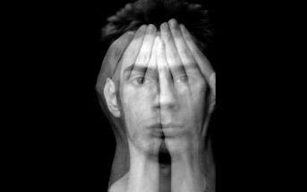 A black and white abstract image showing a man's face blended with overlaying hands covering his eyes. The image creates a double exposure effect, symbolizing vulnerability or introspection against a black background.