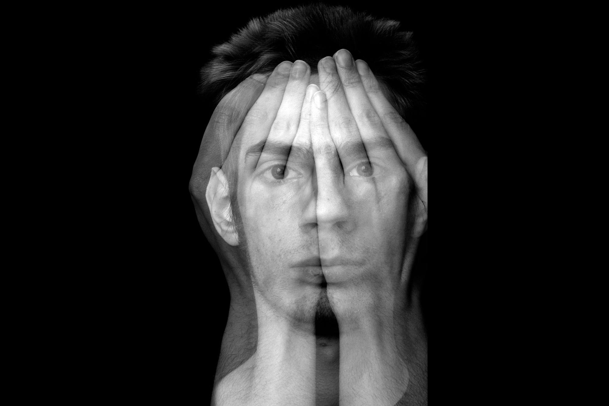 A black and white abstract image showing a man's face blended with overlaying hands covering his eyes. The image creates a double exposure effect, symbolizing vulnerability or introspection against a black background.