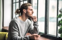 A person with long hair tied back, wearing a gray sweater, sits holding a smartphone and gazes thoughtfully out of a large window. Natural light illuminates the scene, with a plant visible on the windowsill.