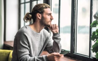 A person with long hair tied back, wearing a gray sweater, sits holding a smartphone and gazes thoughtfully out of a large window. Natural light illuminates the scene, with a plant visible on the windowsill.