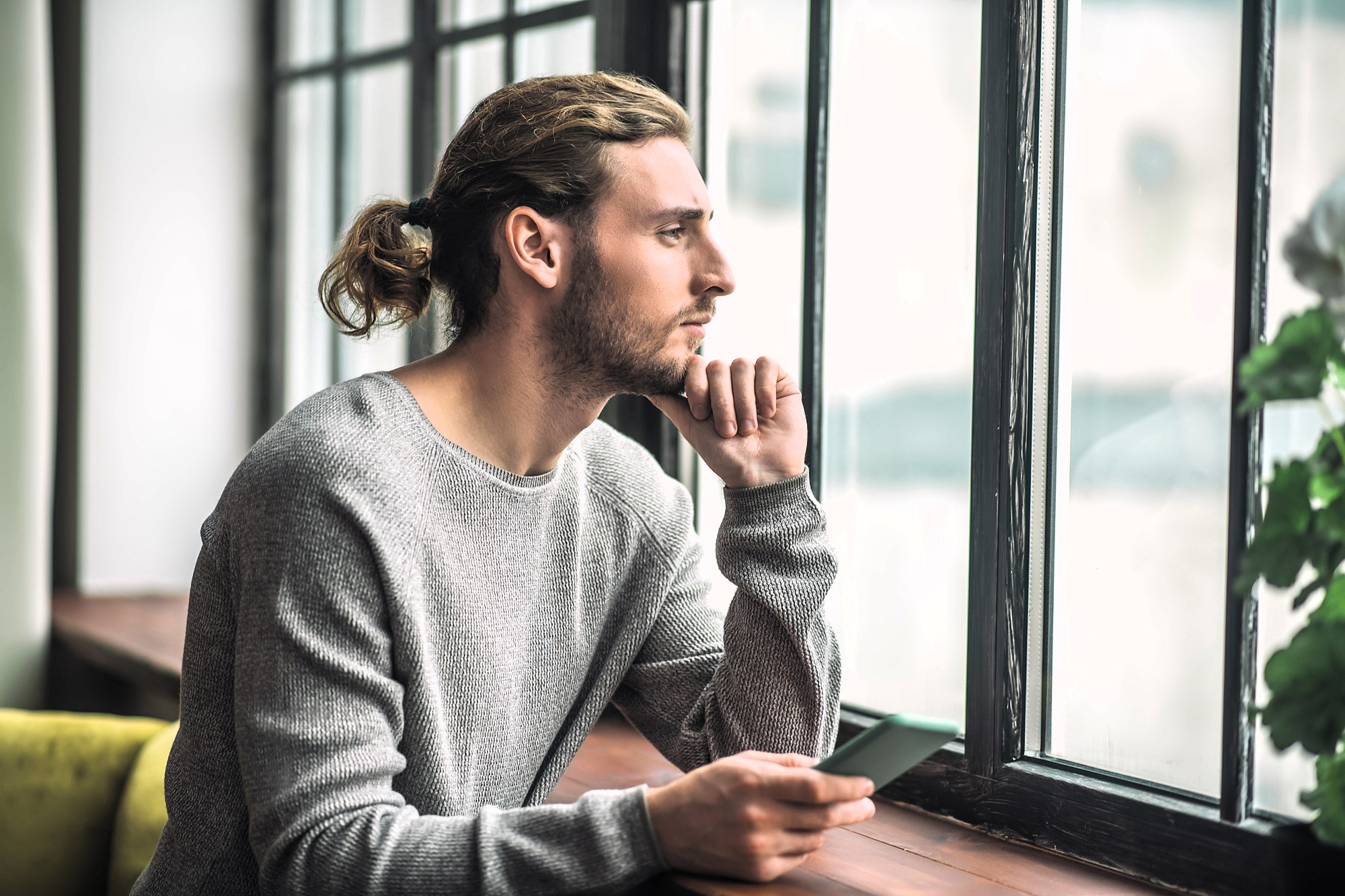 A person with long hair tied back, wearing a gray sweater, sits holding a smartphone and gazes thoughtfully out of a large window. Natural light illuminates the scene, with a plant visible on the windowsill.