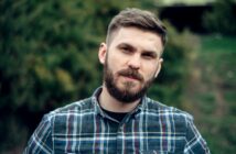 A man with a beard and short hair, wearing a plaid shirt, stands outdoors. The background is green and blurred, suggesting a natural setting. He looks directly at the camera with a neutral expression.