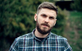 A man with a beard and short hair, wearing a plaid shirt, stands outdoors. The background is green and blurred, suggesting a natural setting. He looks directly at the camera with a neutral expression.