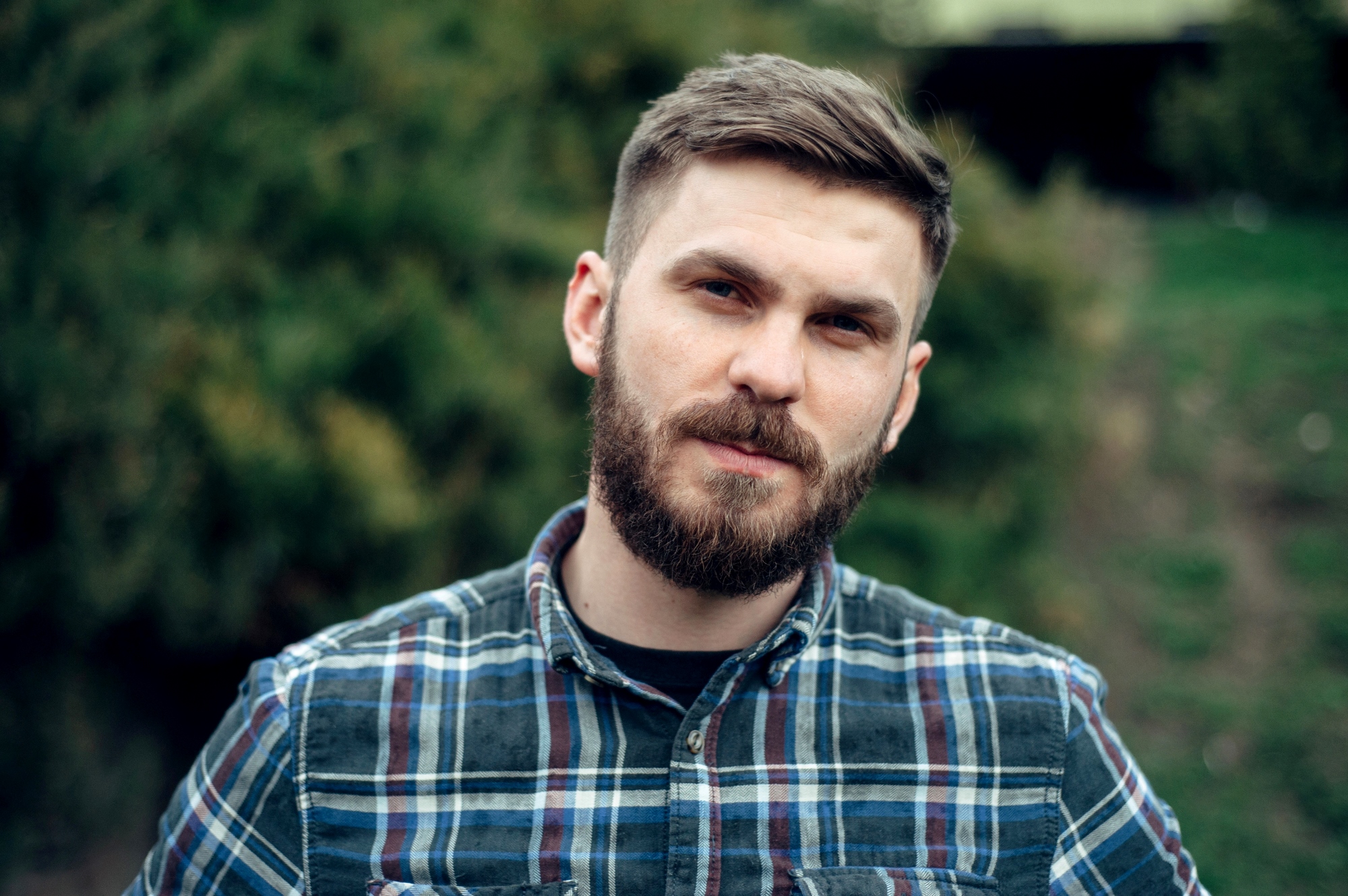 A man with a beard and short hair, wearing a plaid shirt, stands outdoors. The background is green and blurred, suggesting a natural setting. He looks directly at the camera with a neutral expression.