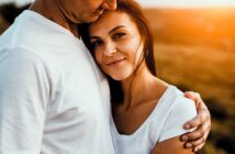 A woman with long brown hair smiles, nestled in the embrace of a man wearing a white shirt. The sun casts a warm glow, creating a serene and tender atmosphere. They both wear white shirts, enhancing the peaceful setting.