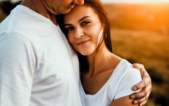 A woman with long brown hair smiles, nestled in the embrace of a man wearing a white shirt. The sun casts a warm glow, creating a serene and tender atmosphere. They both wear white shirts, enhancing the peaceful setting.