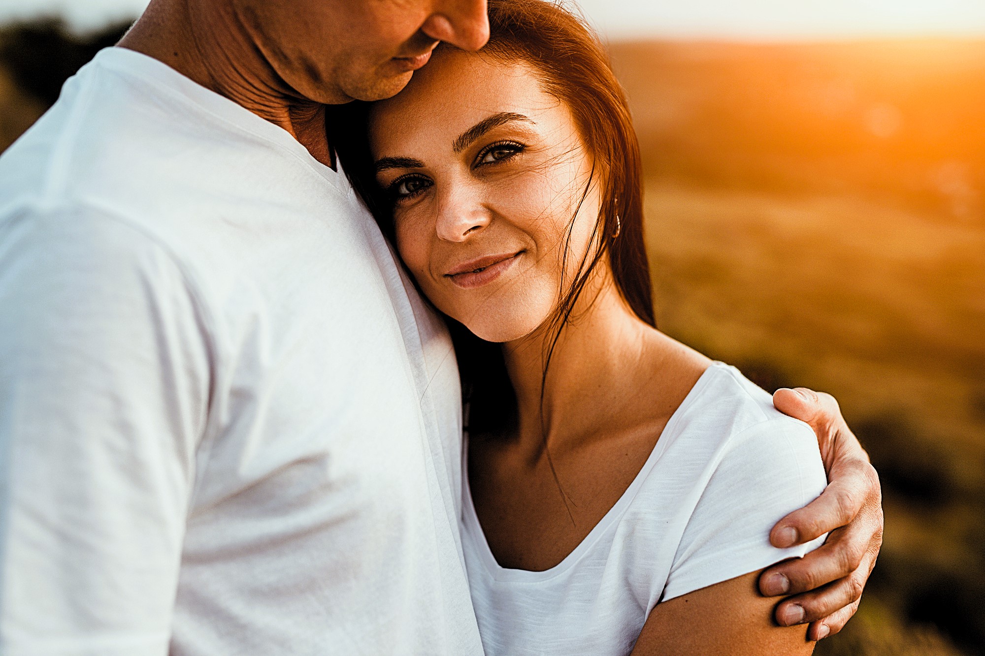 A woman with long brown hair smiles, nestled in the embrace of a man wearing a white shirt. The sun casts a warm glow, creating a serene and tender atmosphere. They both wear white shirts, enhancing the peaceful setting.