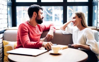 A man and woman sit across from each other in a cozy cafe, engaging in conversation. The man wears a red sweater, and the woman wears a white one. There's a coffee cup, a tablet, and a notebook on the table. Large windows are in the background.