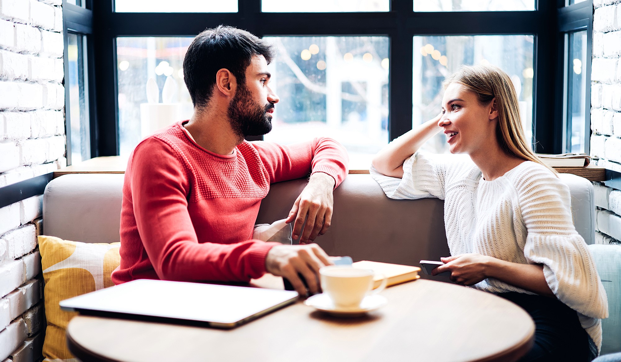 A man and woman sit across from each other in a cozy cafe, engaging in conversation. The man wears a red sweater, and the woman wears a white one. There's a coffee cup, a tablet, and a notebook on the table. Large windows are in the background.