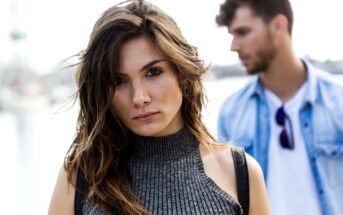 A woman with long brown hair and a gray sleeveless turtleneck looks directly at the camera. In the background, a man with brown hair and a denim shirt stands slightly out of focus. The setting appears to be outdoors.