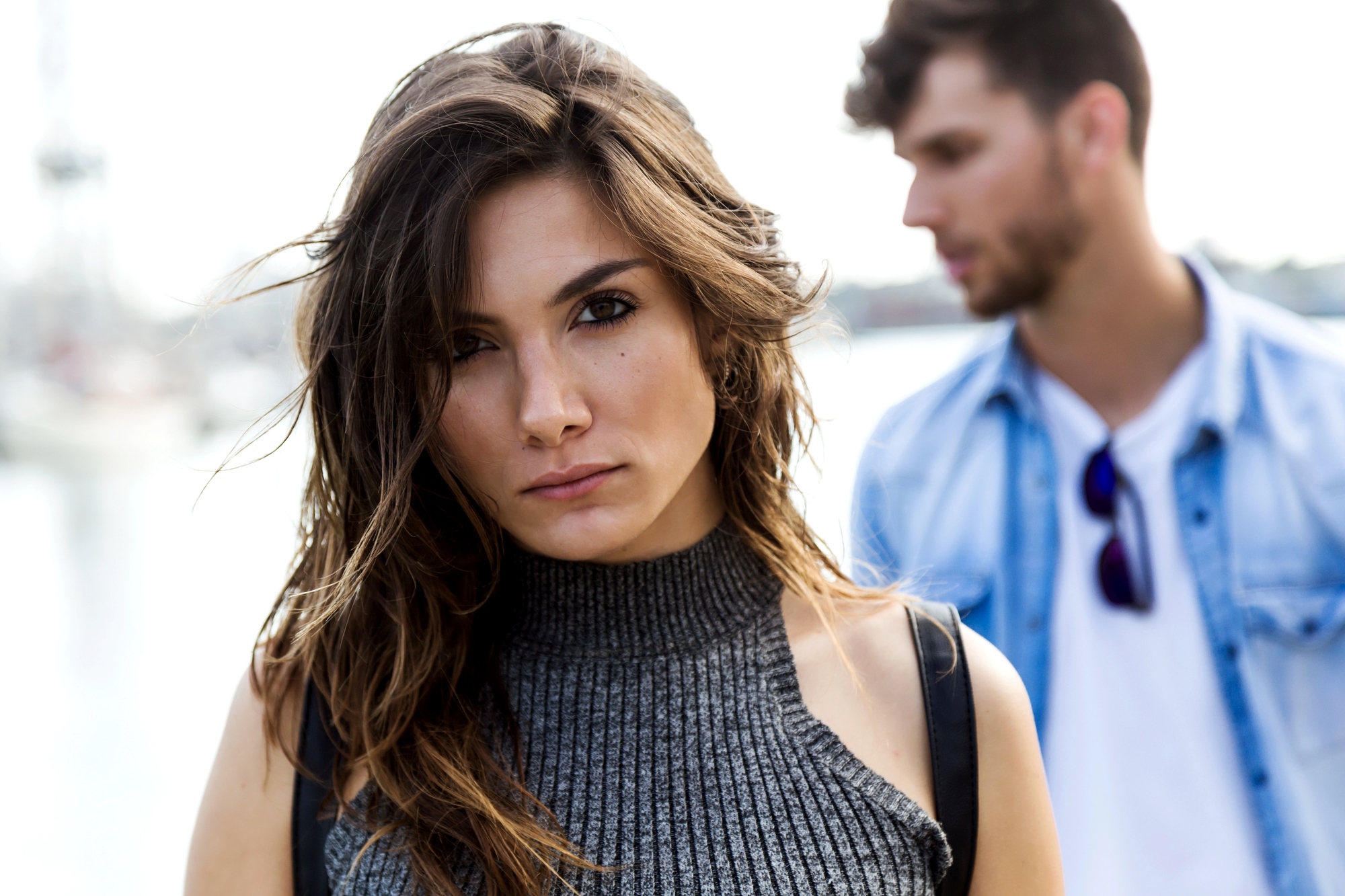 A woman with long brown hair and a gray sleeveless turtleneck looks directly at the camera. In the background, a man with brown hair and a denim shirt stands slightly out of focus. The setting appears to be outdoors.