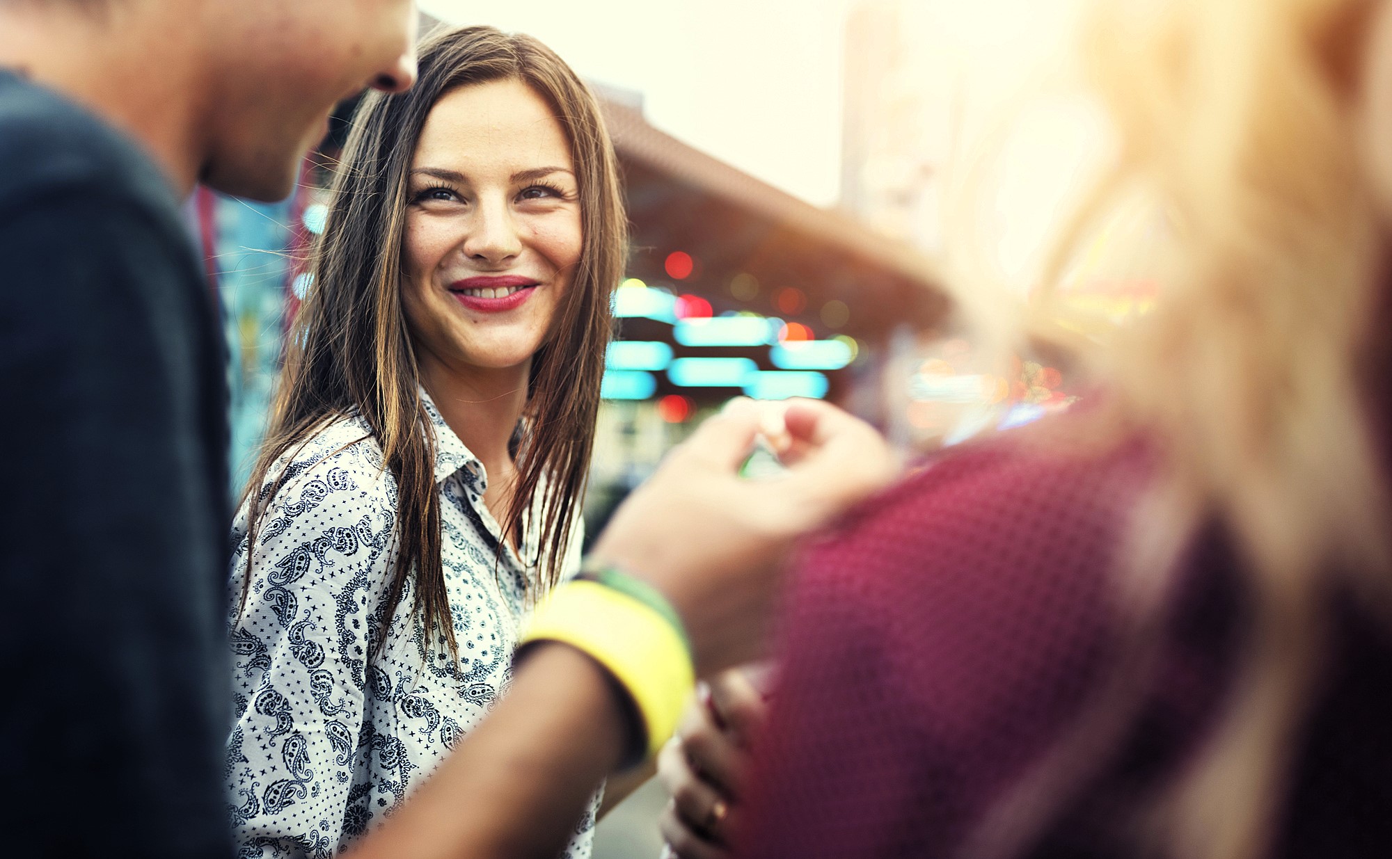 A woman with long brown hair, wearing a paisley-patterned shirt, smiles while outdoors. She is surrounded by other people in a lively, sunlit setting, suggesting a casual social event.