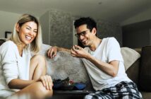 A woman and a man sit on a couch, laughing together. They wear casual clothing, and several bowls of food are on a small table in front of them. The room is warmly lit, and they appear to be enjoying a relaxed and joyful moment.