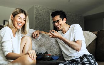 A woman and a man sit on a couch, laughing together. They wear casual clothing, and several bowls of food are on a small table in front of them. The room is warmly lit, and they appear to be enjoying a relaxed and joyful moment.