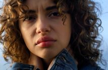 A person with curly hair gazes into the distance, wearing a denim jacket and holding the collar close to their neck. The background is a soft, out-of-focus sky.