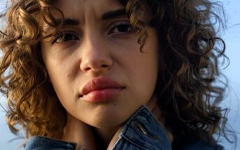 A person with curly hair gazes into the distance, wearing a denim jacket and holding the collar close to their neck. The background is a soft, out-of-focus sky.