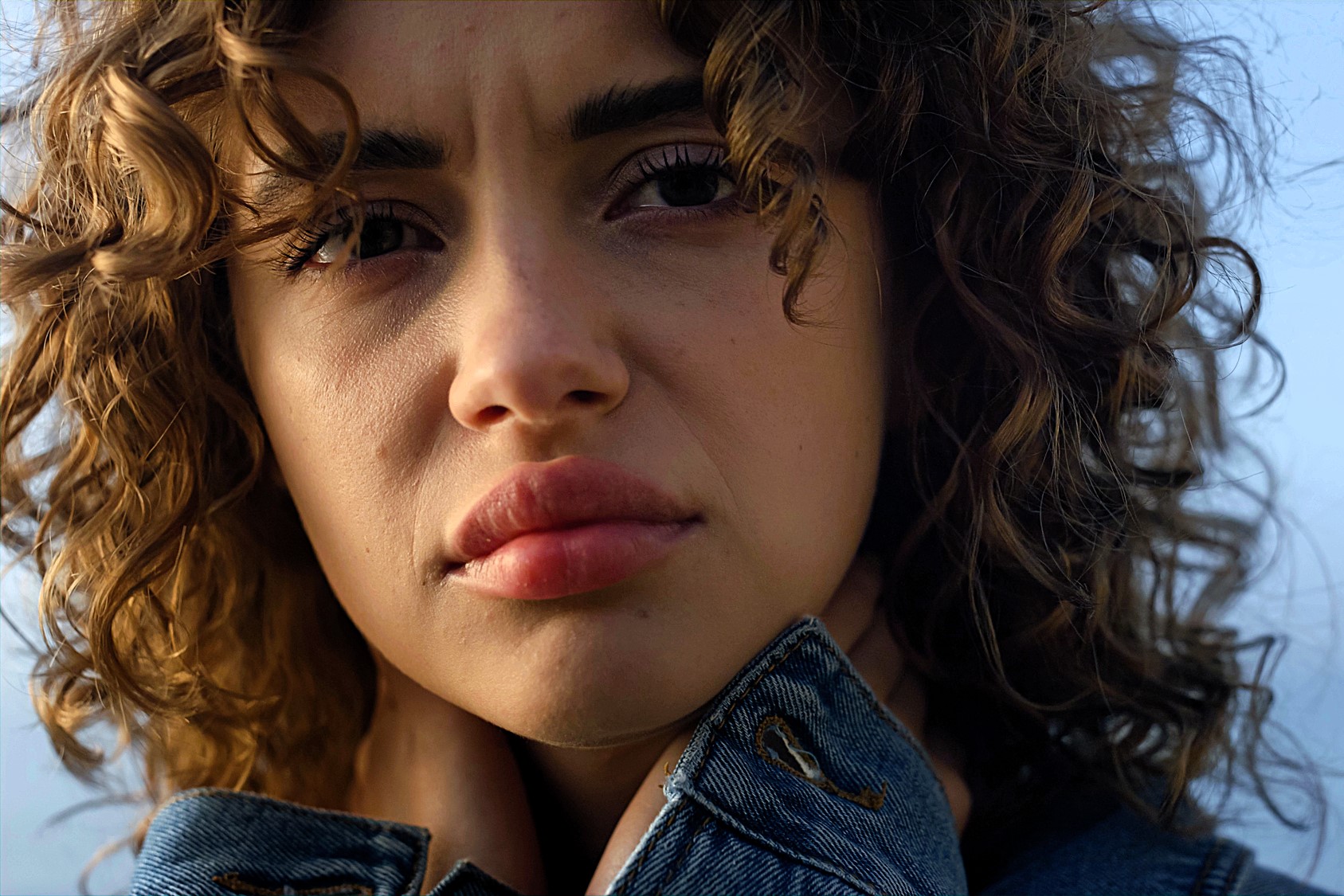 A person with curly hair gazes into the distance, wearing a denim jacket and holding the collar close to their neck. The background is a soft, out-of-focus sky.