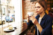 A woman with blonde hair sits at a wooden table by a window, holding a white cup. She is looking intently at the camera. A slice of cake is on a plate in front of her. The background shows a blurred street view through the window.