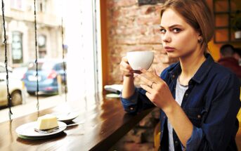 A woman with blonde hair sits at a wooden table by a window, holding a white cup. She is looking intently at the camera. A slice of cake is on a plate in front of her. The background shows a blurred street view through the window.