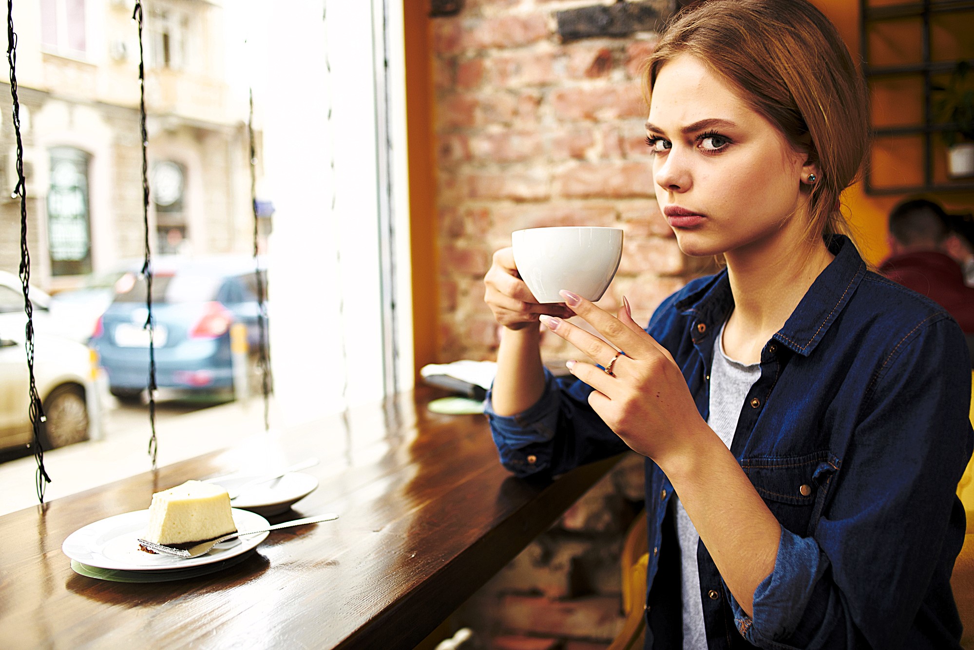 A woman with blonde hair sits at a wooden table by a window, holding a white cup. She is looking intently at the camera. A slice of cake is on a plate in front of her. The background shows a blurred street view through the window.