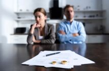 A man and woman sit at a wooden table, appearing distant and thoughtful. In the foreground, divorce papers with a pen and two wedding rings are visible, suggesting a focus on the couple's separation. The background shows a blurred kitchen setting.