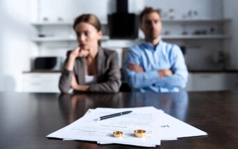 A man and woman sit at a wooden table, appearing distant and thoughtful. In the foreground, divorce papers with a pen and two wedding rings are visible, suggesting a focus on the couple's separation. The background shows a blurred kitchen setting.