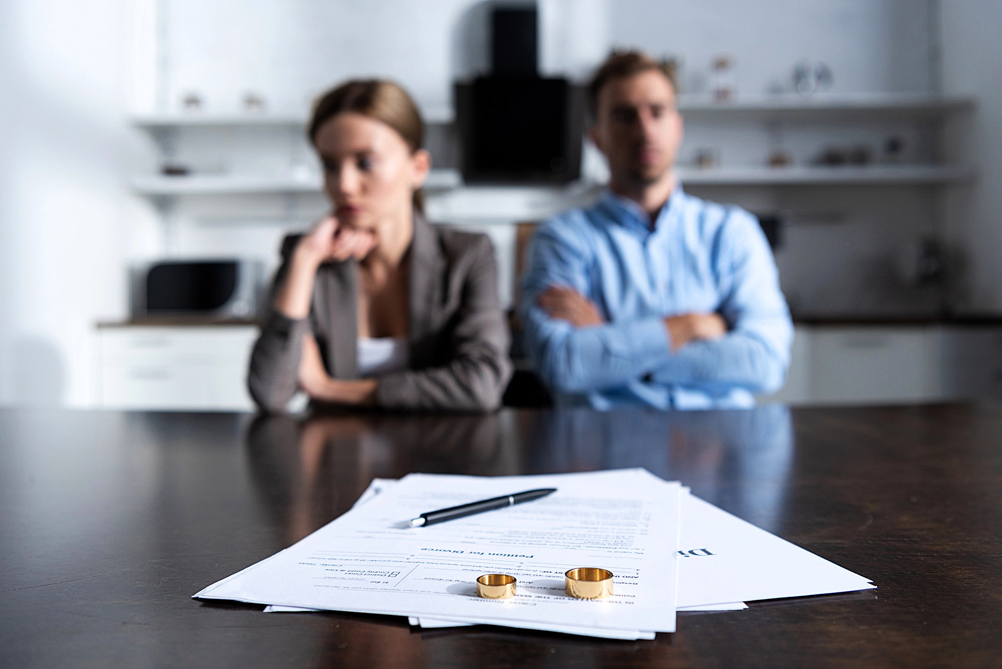 A man and woman sit at a wooden table, appearing distant and thoughtful. In the foreground, divorce papers with a pen and two wedding rings are visible, suggesting a focus on the couple's separation. The background shows a blurred kitchen setting.