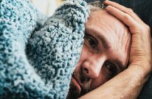 A person lies on a bed, partially covered by a fluffy blue blanket. They appear contemplative and are resting their head on a pillow with one hand on their forehead. The mood suggests introspection or concern.