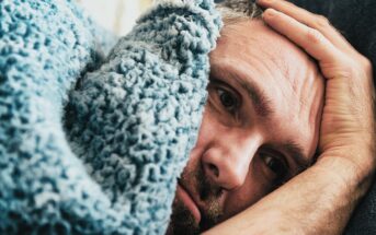 A person lies on a bed, partially covered by a fluffy blue blanket. They appear contemplative and are resting their head on a pillow with one hand on their forehead. The mood suggests introspection or concern.