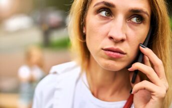 A woman with blonde hair looks concerned as she holds a phone to her ear. She is wearing a white shirt and has dark nails. The background is blurred, with an indistinct figure in the distance.