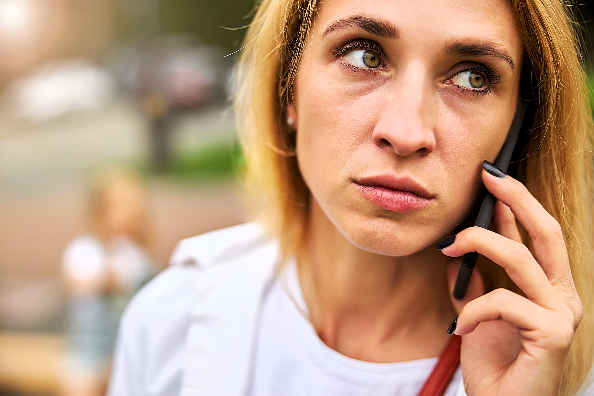 A woman with blonde hair looks concerned as she holds a phone to her ear. She is wearing a white shirt and has dark nails. The background is blurred, with an indistinct figure in the distance.