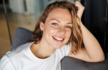 A person with shoulder-length hair smiles warmly, leaning on a gray couch. They are wearing a light-colored t-shirt, and the background features blurred indoor elements.