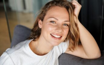 A person with shoulder-length hair smiles warmly, leaning on a gray couch. They are wearing a light-colored t-shirt, and the background features blurred indoor elements.